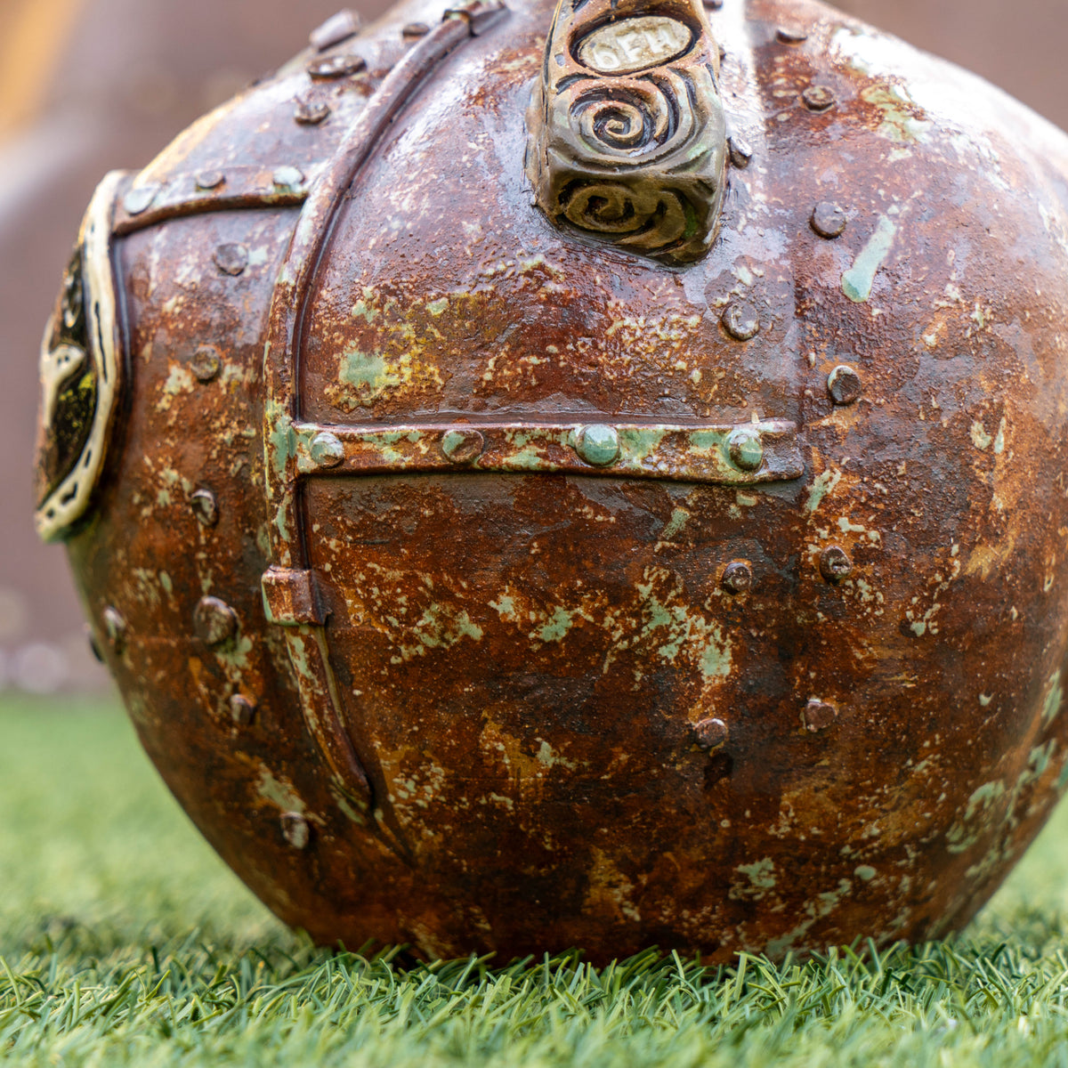 A close-up of the Steampunk Punkin Ceramic Growler by Romanick Pottery—a rusted, riveted metal-like sphere with green patina—rests on vivid green grass. The growler features bolts, seams, and a decorative metallic pattern on its surface, showcasing the sturdy craftsmanship characteristic of Romanick Pottery’s designs.