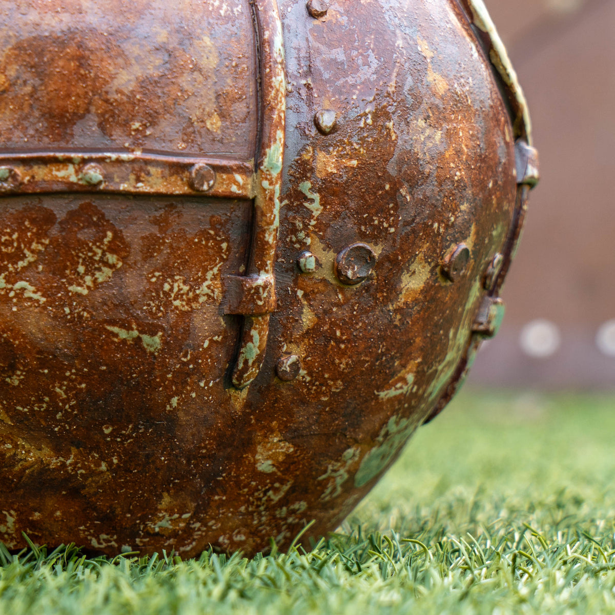 A close-up of a rusty, riveted metal object resting on green grass, its weathered textures and hints of green corrosion evoke the raw aesthetic and distinctive character found in Romanick Pottery’s Steampunk Punkin Ceramic Growler.