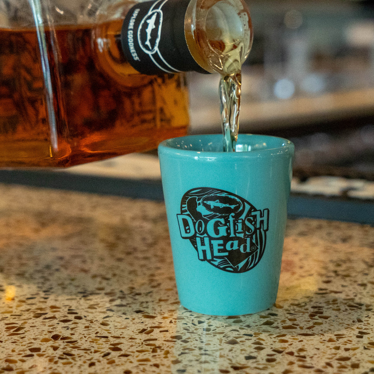 Teal shot glass sitting on speckled table having spirits  poured into it.  Logo says 'dogfish head'