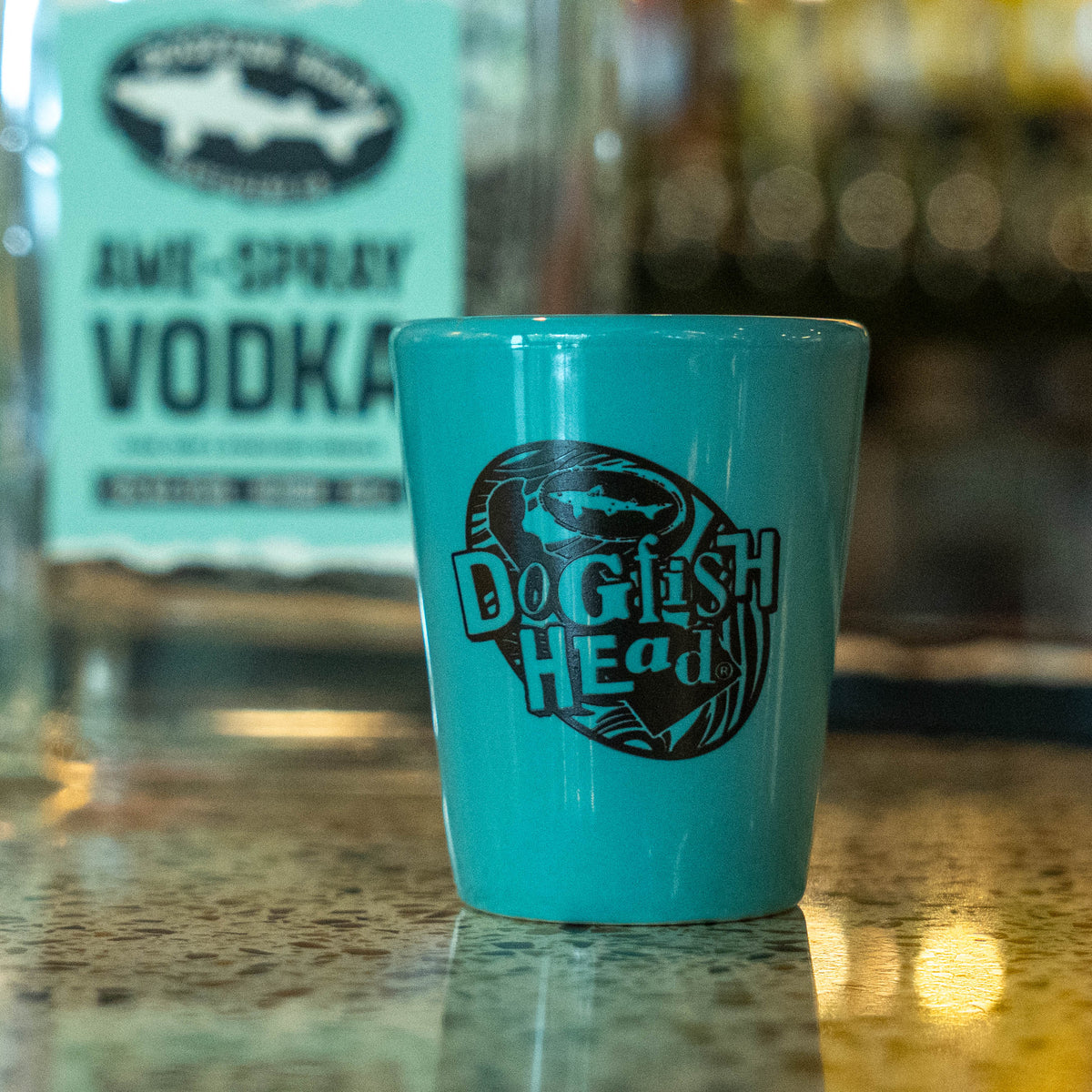 A teal handcrafted Grandstand Glassware & Apparel Ceramic Shot Glass with a black shark logo sits on a speckled bar top in front of a bottle of vodka.