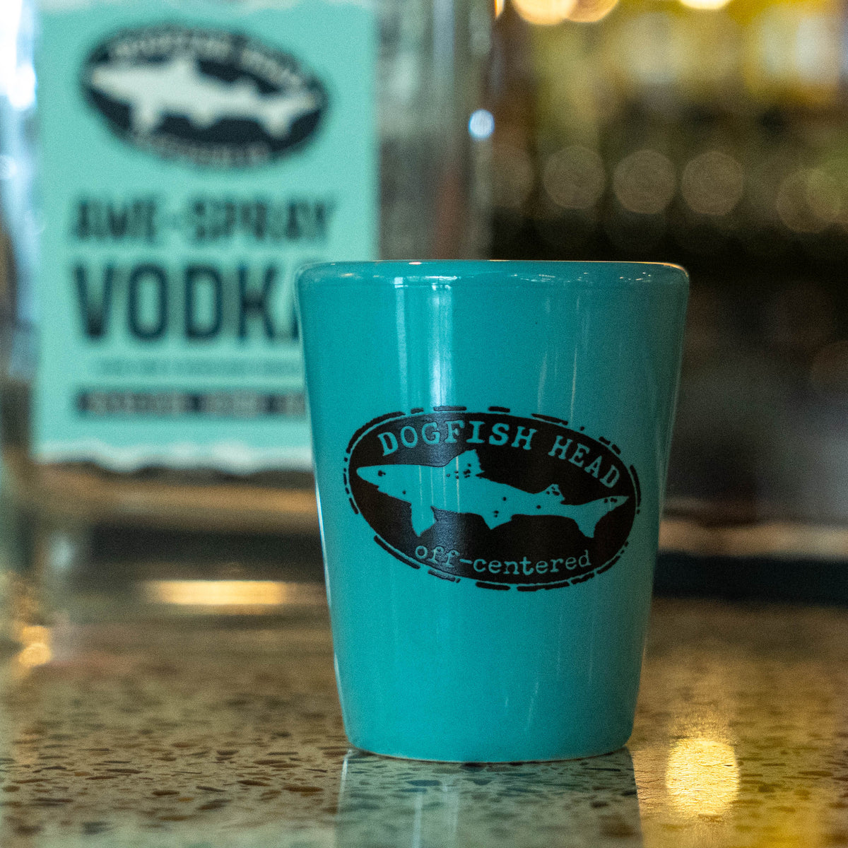Handcrafted teal Ceramic Shot Glass featuring a black logo with a shark silhouette and the text Dogfish Head off-centered, sitting on a speckled bar top.