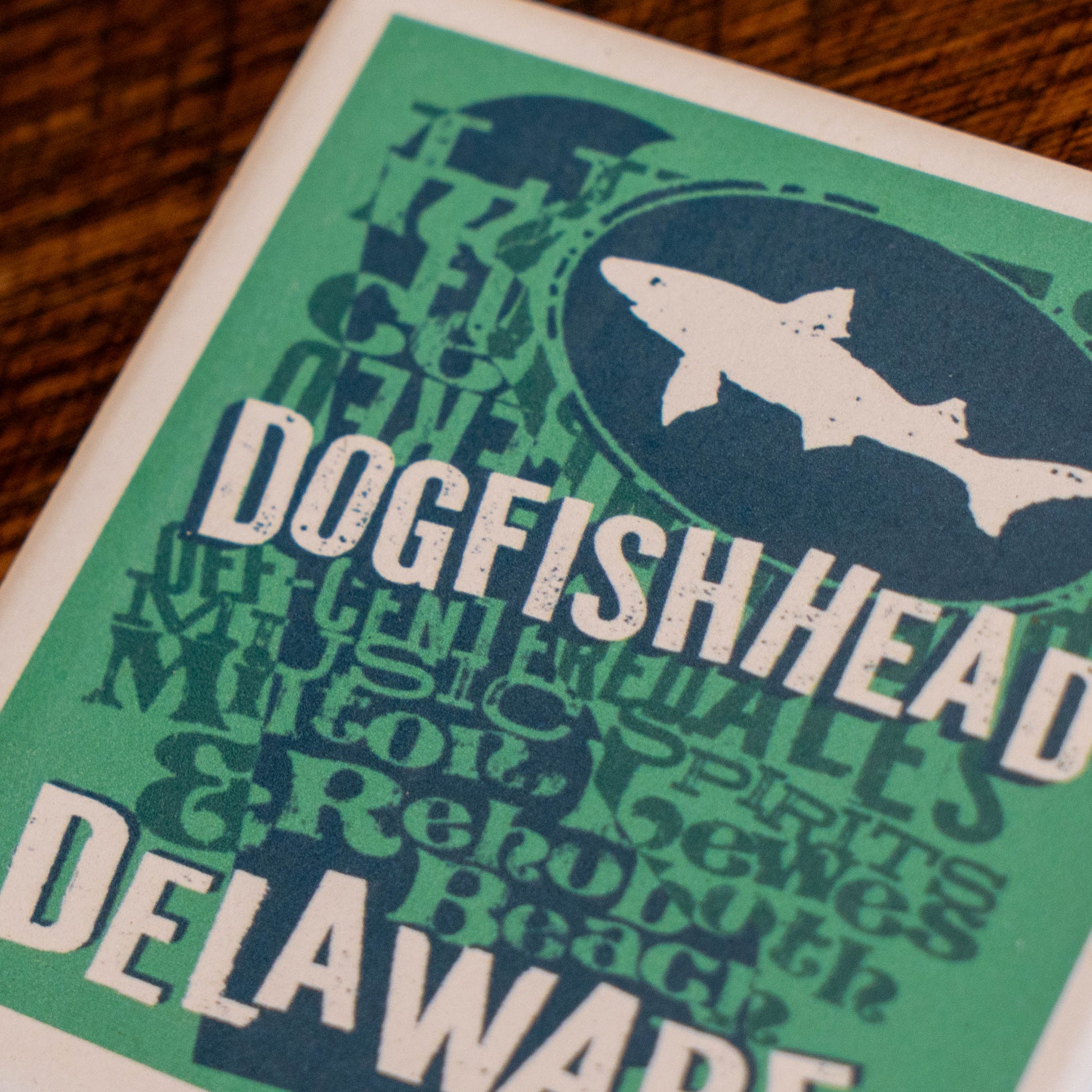 A close-up of a printed coaster featuring a green and navy design with the words 'Dogfish Head' and 'Delaware' plus a white shark silhouette, placed on a wooden surface.