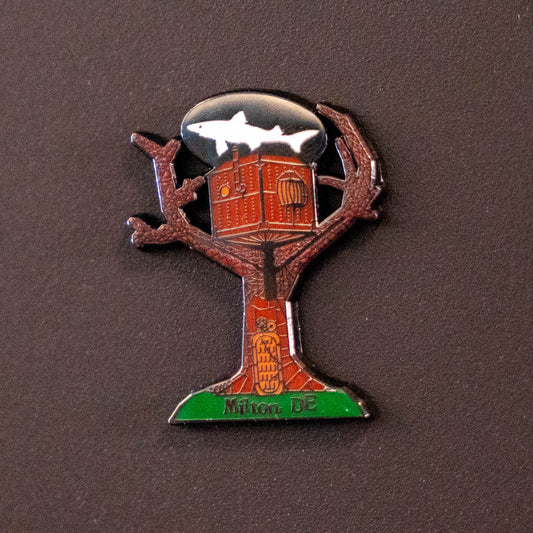 Treehouse Enamel Magnet shaped like a tree with a house in its branches, a white shark silhouette at the top, and the text Milton DE at the base.