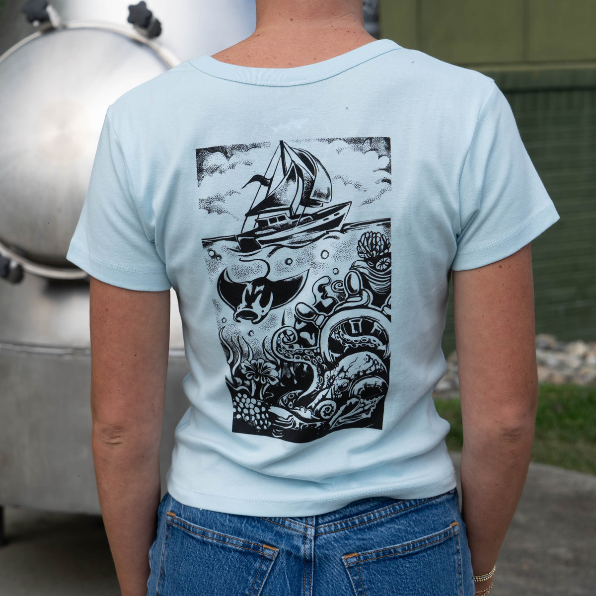W Light Blue Hull and Helm Baby Tee Shirt in light blue, featuring a back graphic of a sailboat on the ocean surface above an underwater scene with a manta ray, an octopus, and coral.