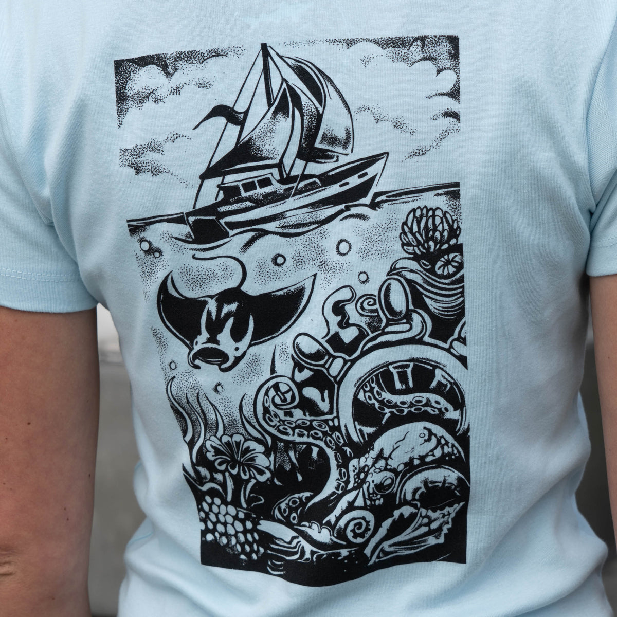W Light Blue Hull and Helm Baby Tee in light blue featuring a black ink illustration of a sailboat on the water with a stingray and octopus swimming below the surface.
