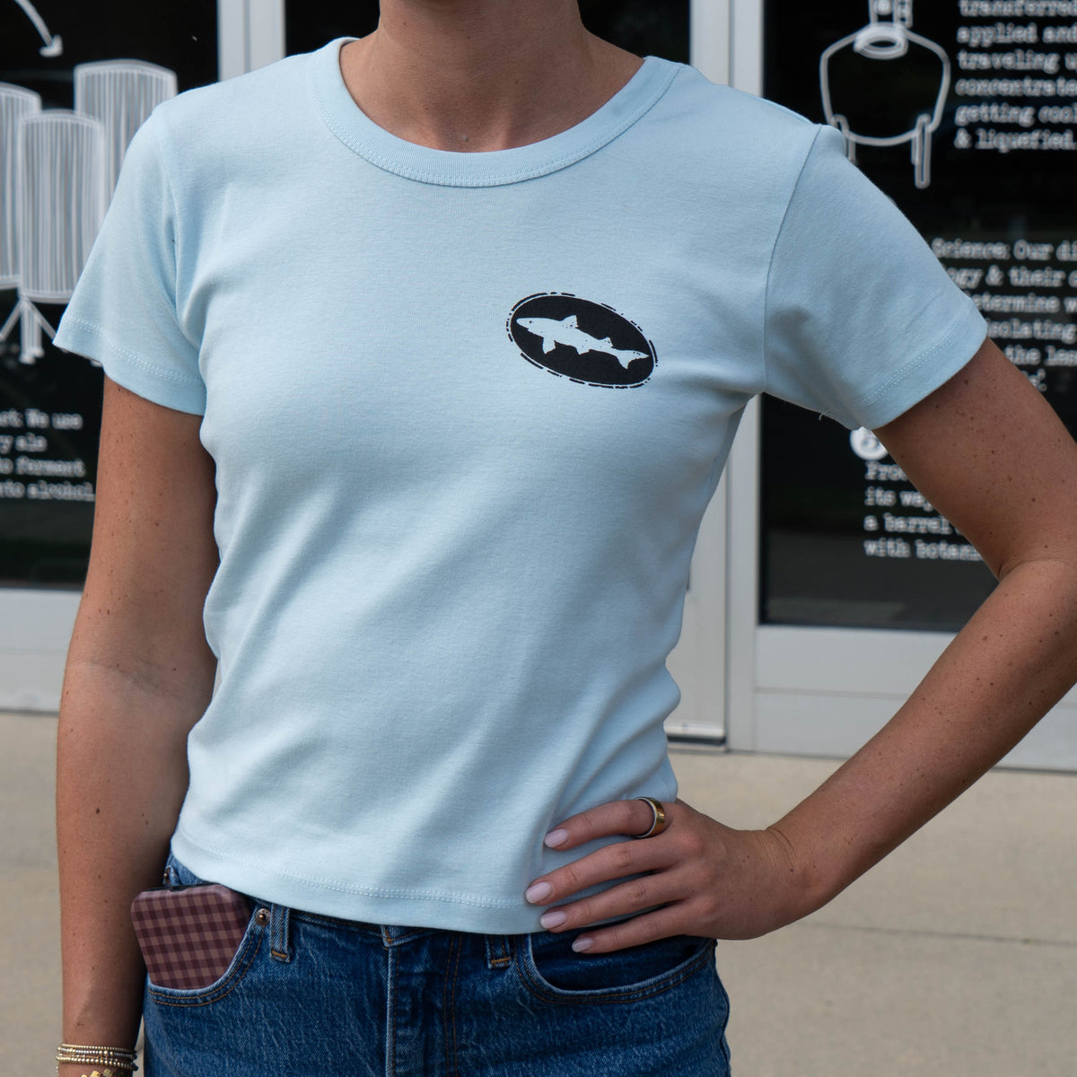 Light Blue short sleeve tee on female.  Black logo on left chest with blue jeans.