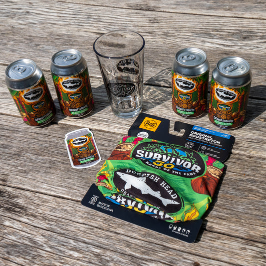 The Dogfish Head x Survivor 50 Bundle by Grandstand Glassware & Apparel—with four vibrant beer cans, a pint glass, sticker, and exclusive Survivor Buff neck gaiter—is displayed on a sunlit wooden surface.