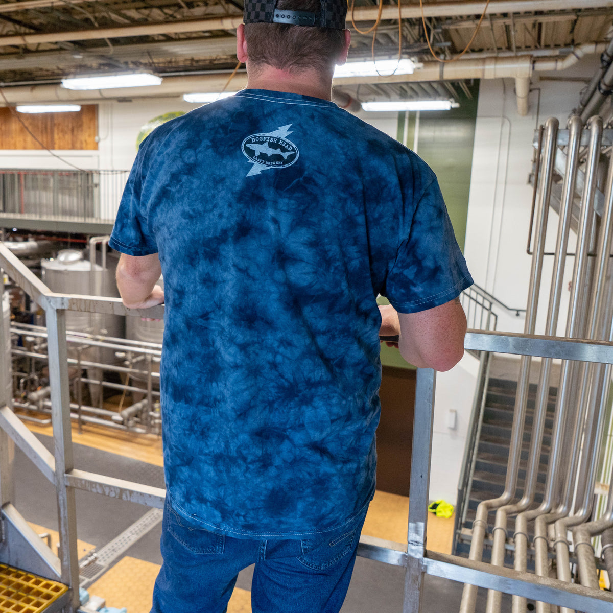 A person seen from behind, wearing a navy blue Dogfish Head x Grateful Dead Navy Tie Dye Short Sleeve Tee Shirt with a white brewery logo on the upper back, standing on a metal catwalk in a brewery.