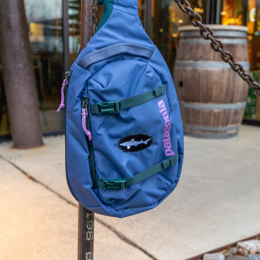 The Patagonia Atom Sling 8L, a blue sling bag with a pink and black logo, hangs outdoors on a metal pole near a chain and barrel. Made from recycled polyester, this ergonomic harness bag is both stylish and sustainable.