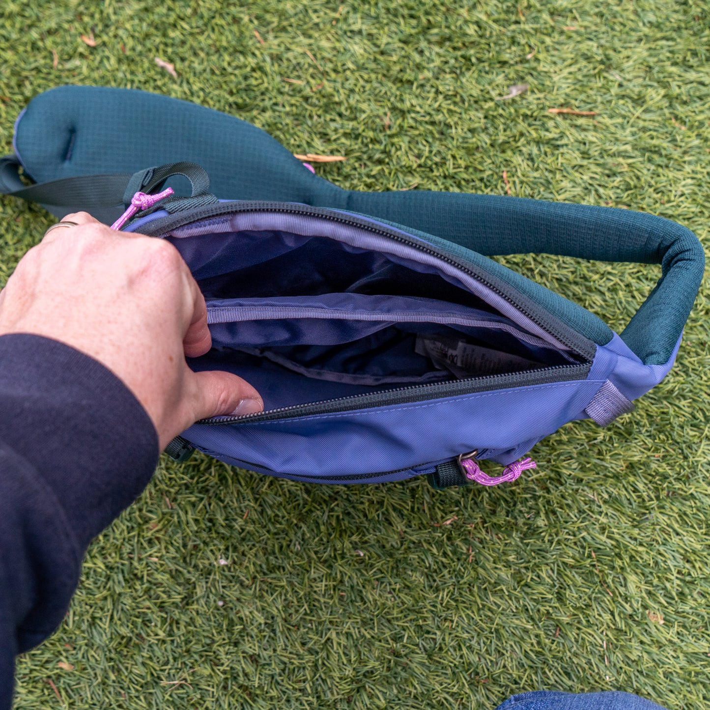 A person stands on green grass holding open a Patagonia Atom Sling 8L, showing its multiple interior compartments.