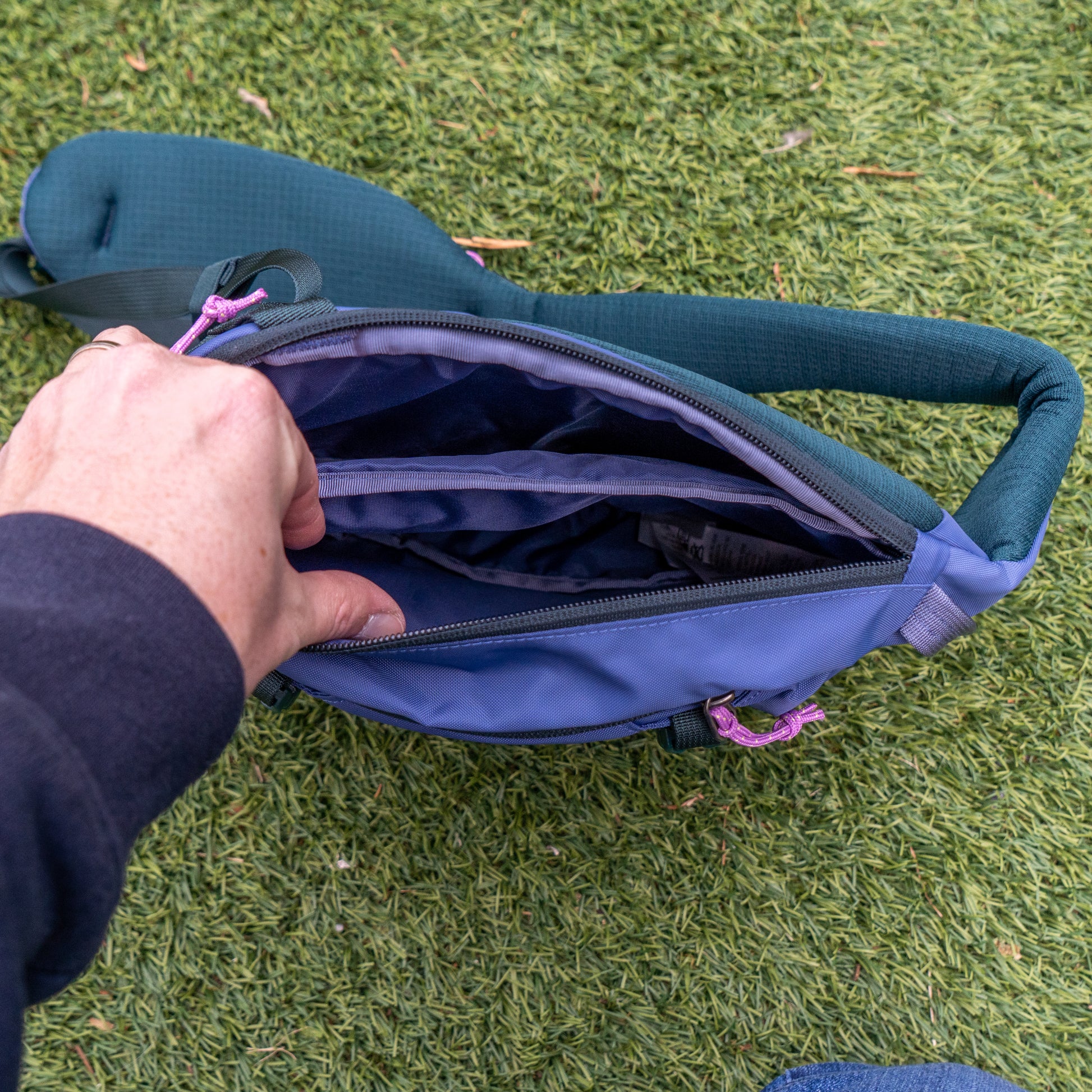 A person stands on green grass holding open a Patagonia Atom Sling 8L, showing its multiple interior compartments.
