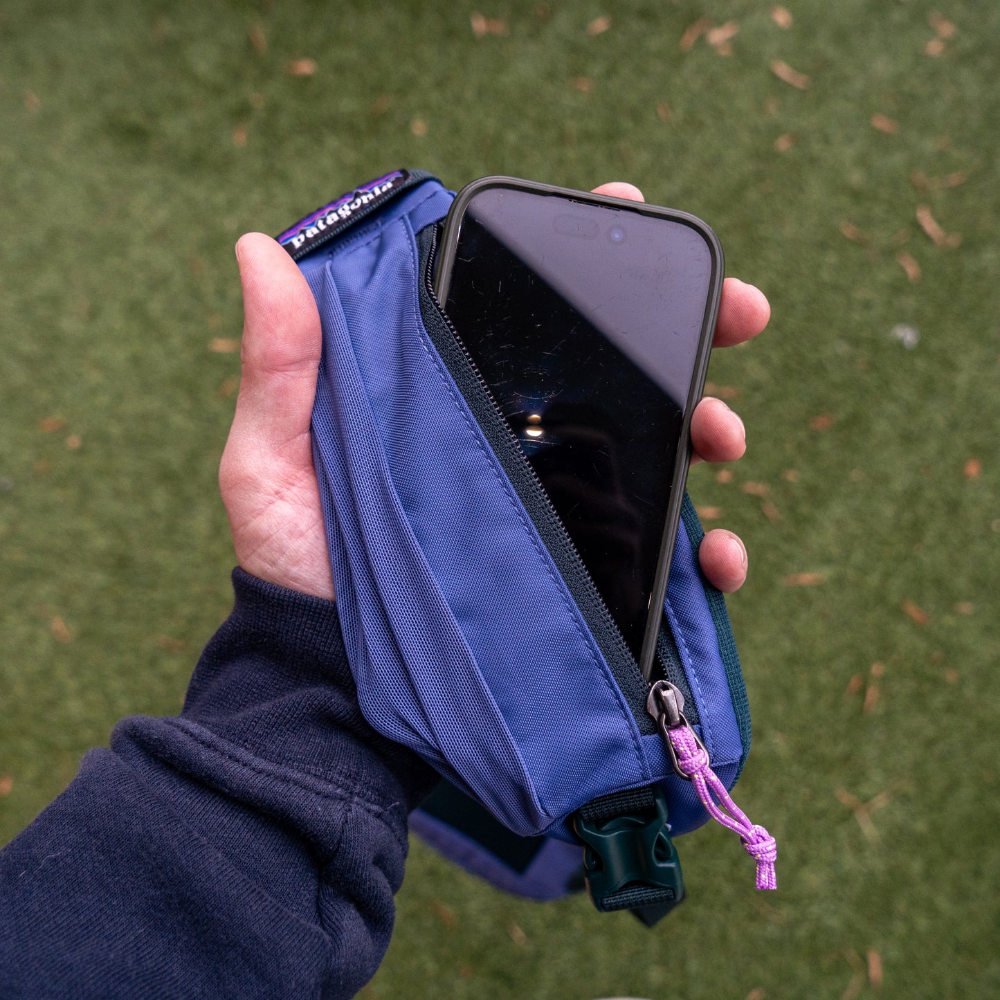 A hand holds the Patagonia Atom Sling 8L, a blue recycled polyester bag with a purple zipper pull and brand tag, partially open to show a phone inside. The sling rests on green grass, combining eco-friendly style with convenience.