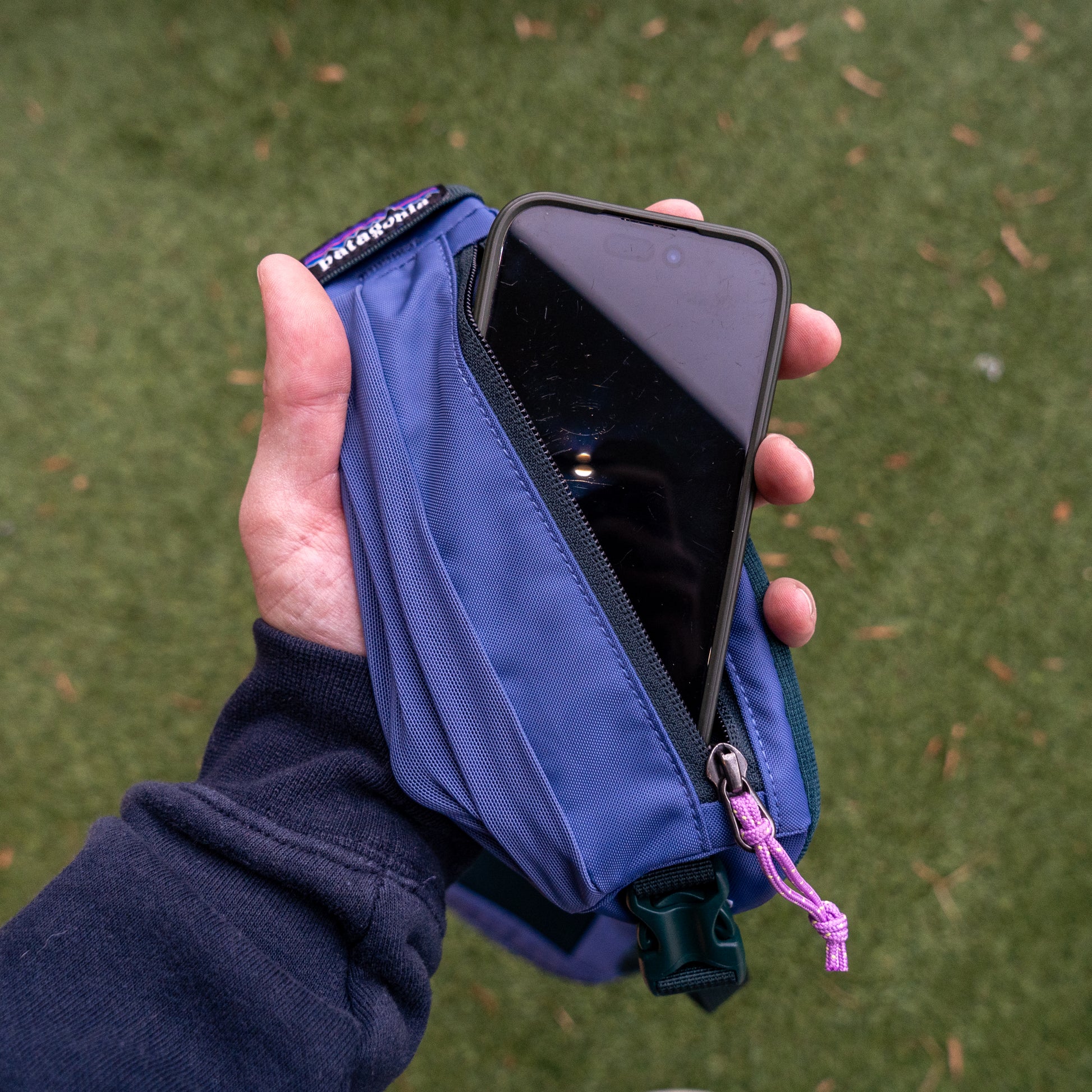 A hand holds the Patagonia Atom Sling 8L, a blue recycled polyester bag with a purple zipper pull and brand tag, partially open to show a phone inside. The sling rests on green grass, combining eco-friendly style with convenience.