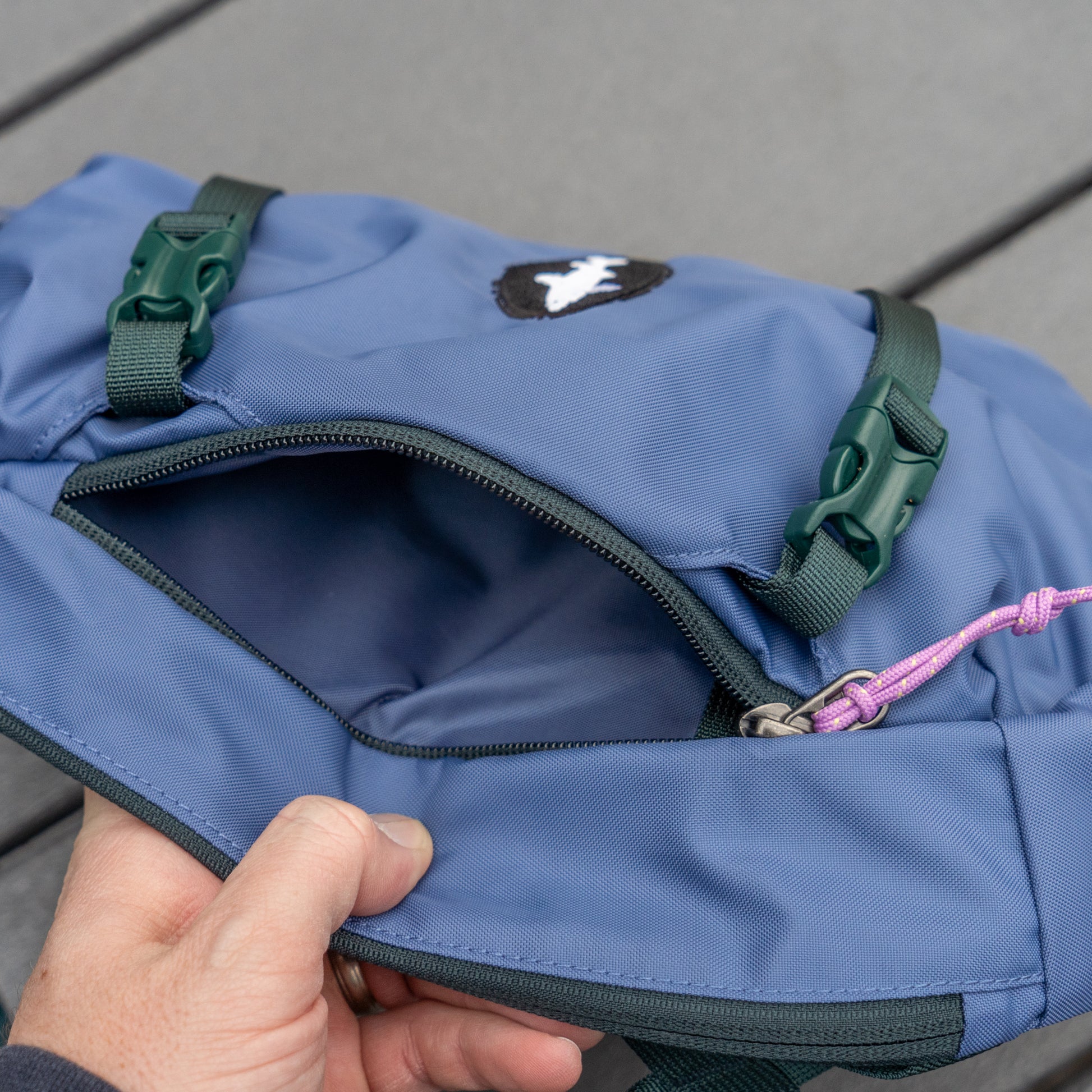 A hand opens the front zippered pocket of the blue Patagonia Atom Sling 8L, made from recycled polyester with green buckles and a pink zipper pull, revealing its empty interior. The Patagonia backpack rests on a wooden surface.