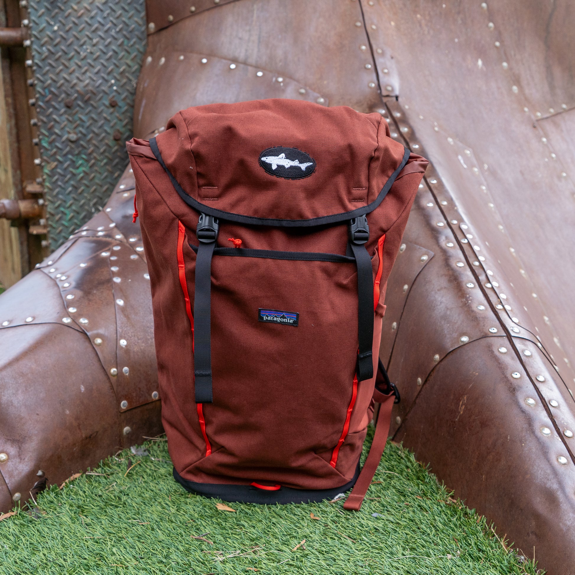 The Patagonia Fieldsmith Lid Pack, an organic cotton backpack in brown and red with a fish-shaped logo, stands upright on artificial grass before a weathered metal slide.