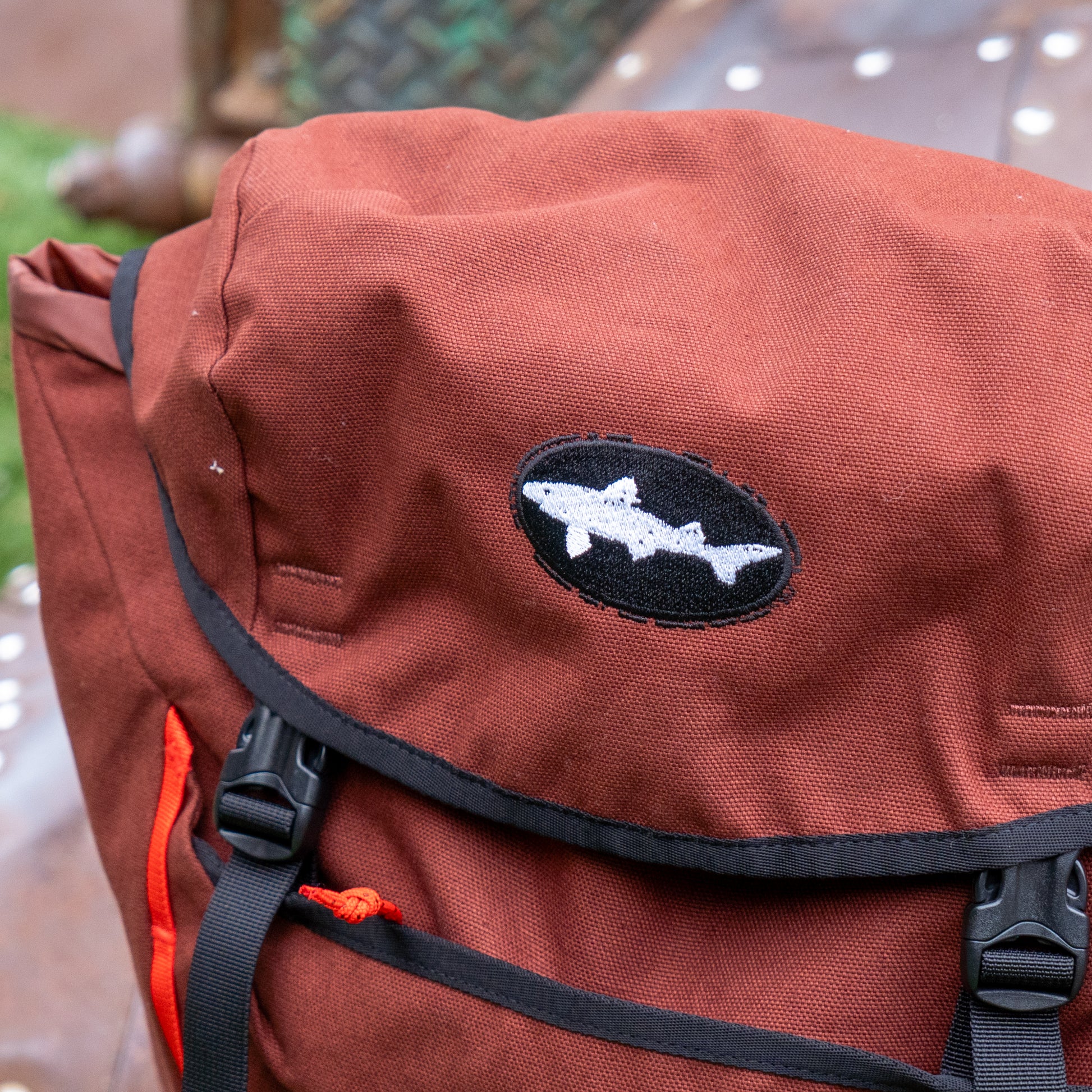 A close-up of Patagonia's Fieldsmith Lid Pack, an organic cotton backpack in rust. It features a black oval patch with a white shark design, water-resistant finish, black trim and clips, and an orange side strap.