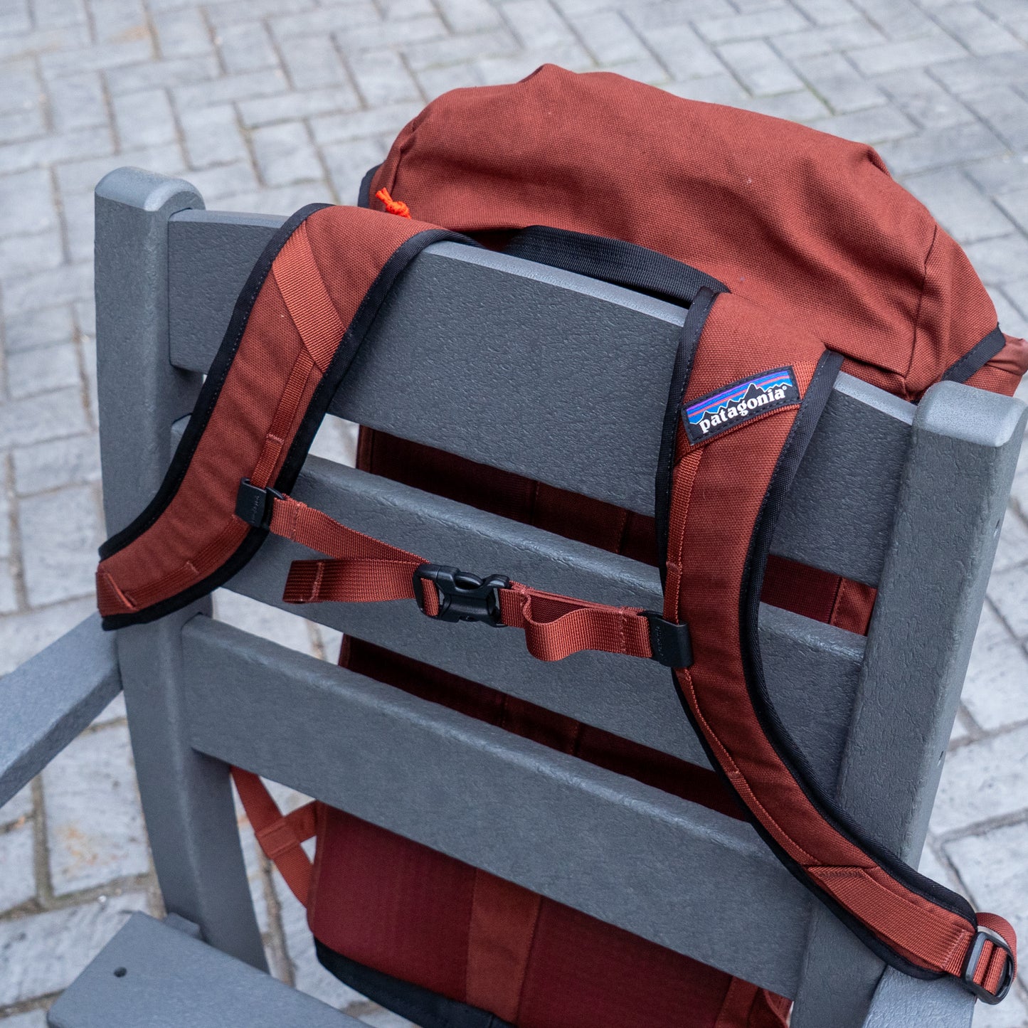 A rust-colored Patagonia Fieldsmith Lid Pack is draped over the back of a gray outdoor chair, showing its shoulder straps and chest buckle. The chair rests on a stone-paved surface.