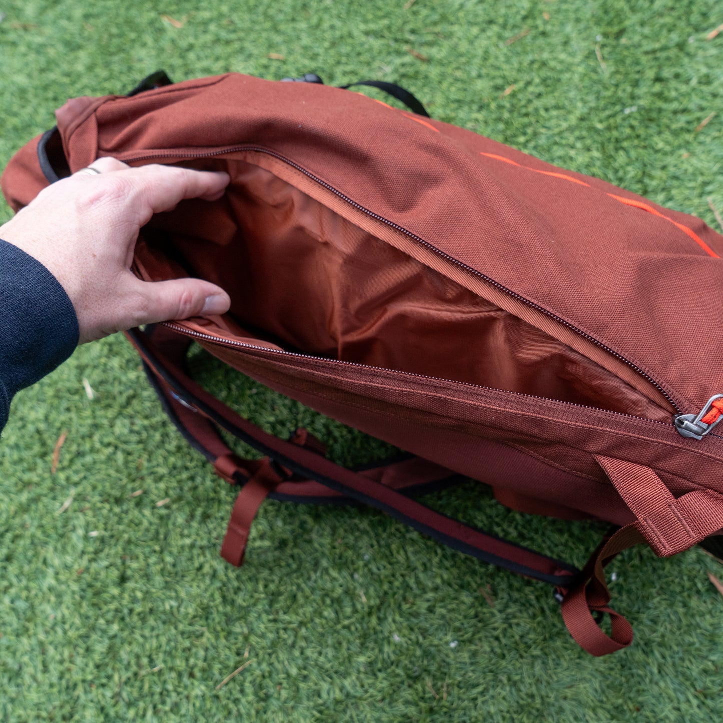 A hand opens the main zipper of the Patagonia Fieldsmith Lid Pack by Patagonia, showing its empty interior. The water-resistant brown organic cotton backpack rests on green grass.