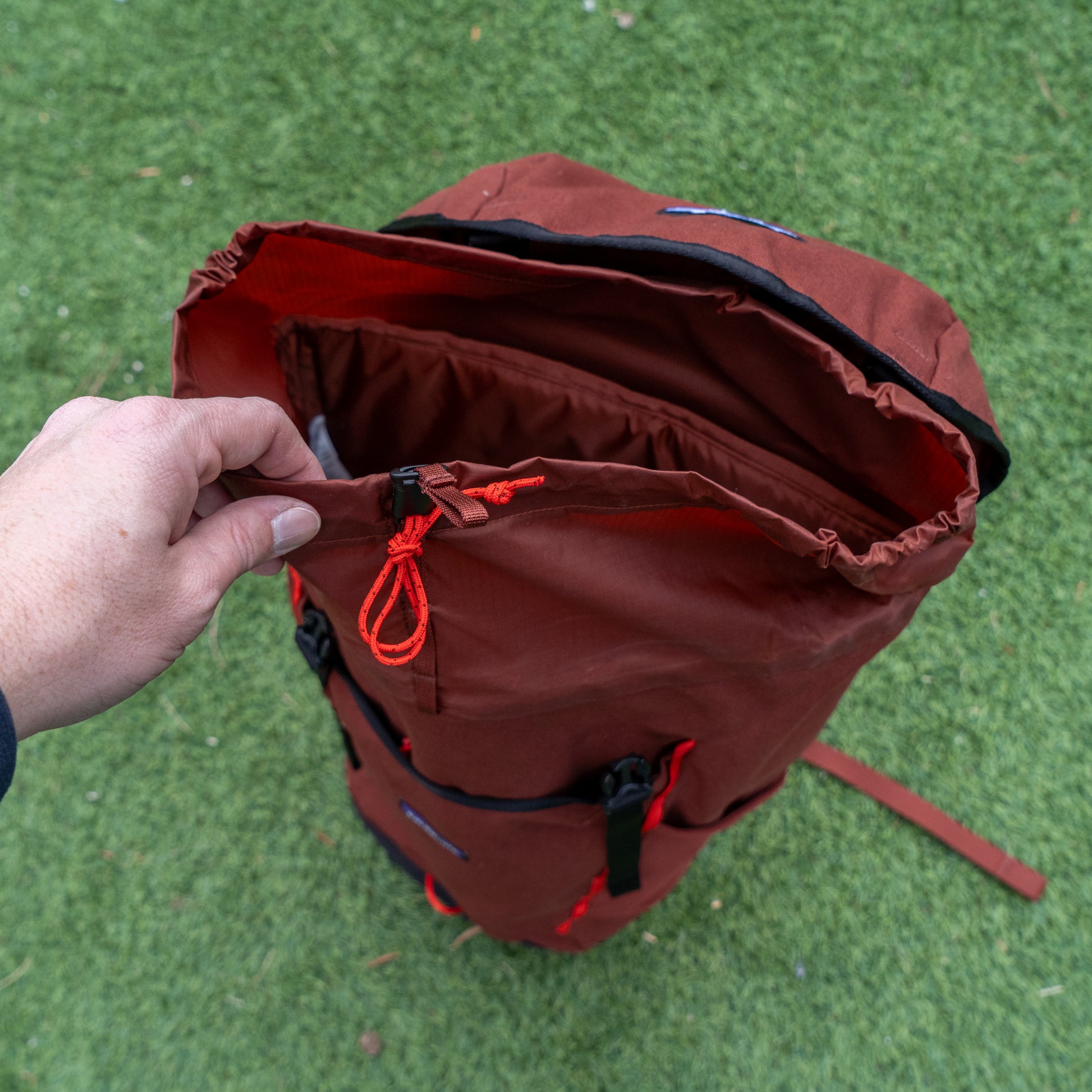 A person’s hand opens the top zipper of a red Patagonia Fieldsmith Lid Pack by Patagonia, which stands upright on green grass.