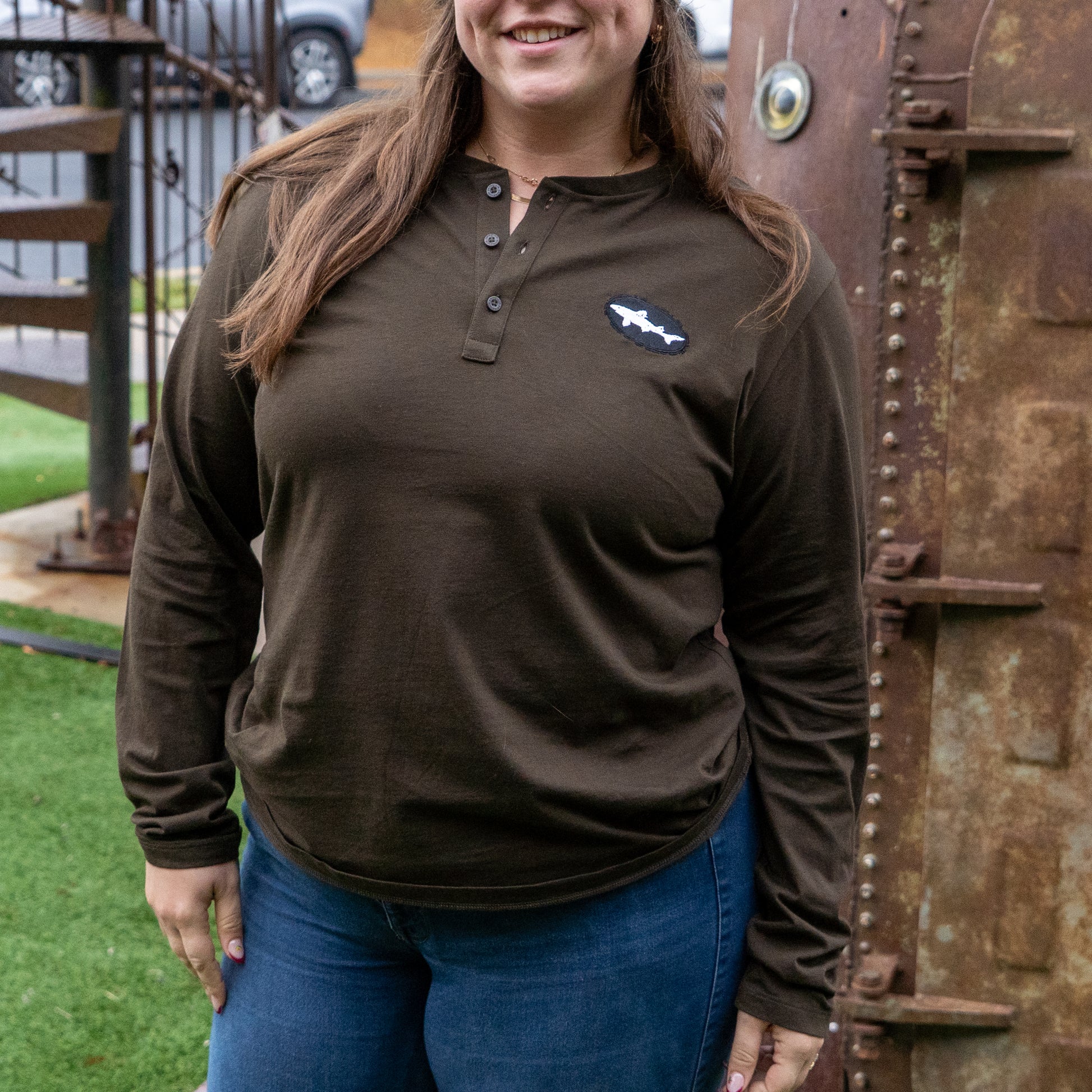 A woman stands smiling on green artificial grass, wearing a Patagonia M Brown Long Sleeve Henley with a small white logo on the left chest. A rusty metal structure is in the background.