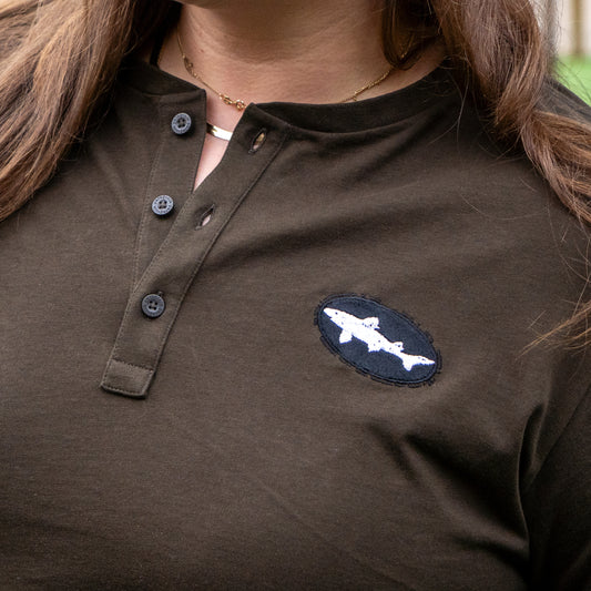 A person with long brown hair wears a Patagonia M Brown Long Sleeve Henley made from regenerative organic cotton, featuring an embroidered white shark patch on the left chest, button-up collar, and a gold necklace.