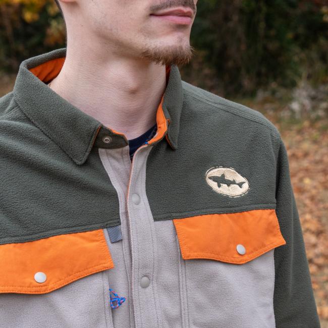 A man wears the Patagonia M Grey Two Pocket Button Down shirt, featuring an olive green and beige design with orange accents and a fish emblem patch, made from recycled polyester microfleece, outdoors among greenery and fallen leaves.