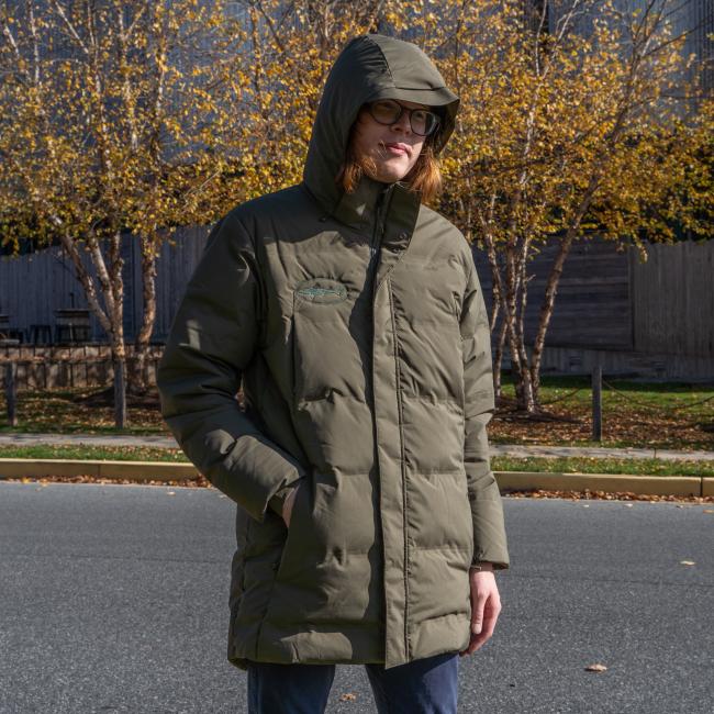 A person in the Patagonia M Olive Green Glacier Parka stands outdoors on a paved street, with autumn trees and a wooden fence in the background.