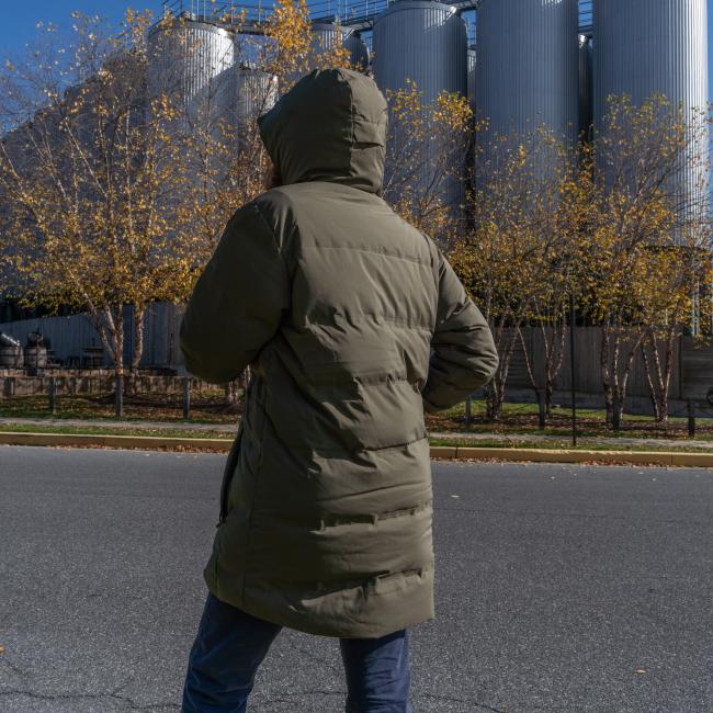 A person in a Patagonia M Olive Green Glacier Parka stands on a street, facing away, with leafless trees and large industrial silos in the background.