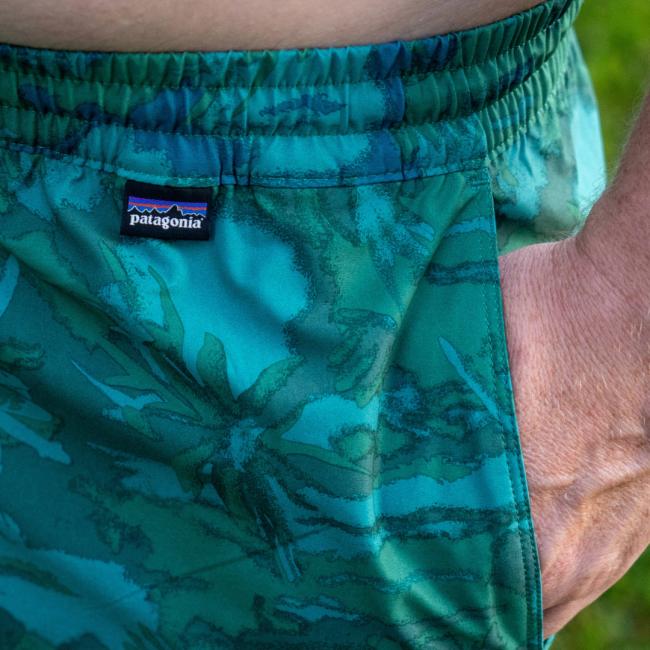 Close-up of a person wearing green patterned Patagonia M Palm Tree Hydropeak Swim Trunks with their right hand in the pocket. The quick-drying trunks feature the Patagonia logo on the waistband.