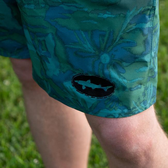 A close-up of a person wearing Patagonia M Palm Tree Hydropeak Swim Trunks, featuring a green and blue palm tree print, standing on grass.