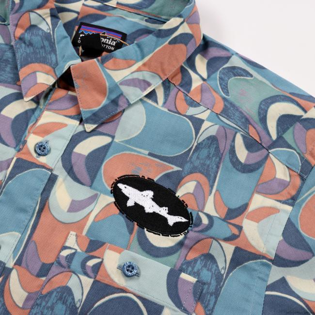 A close-up of the Patagonia Multi Geo Shape Button Up, an organic cotton shirt with an abstract blue, peach, and white geo print. Features a chest pocket, a Dogfish Head patch with two white fish on black, and a visible Patagonia label.