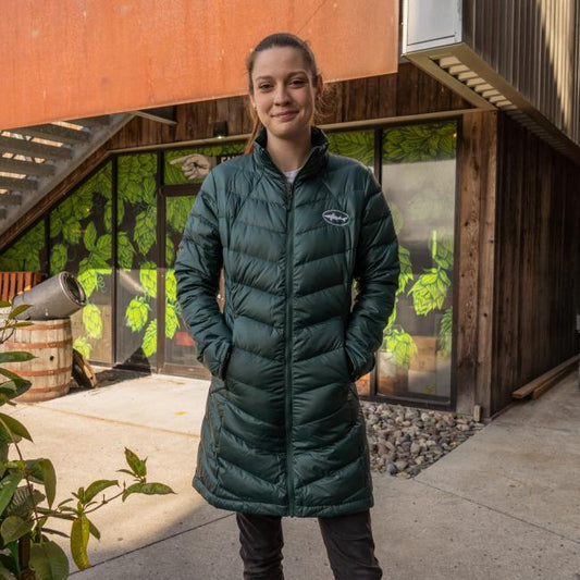 A person wearing the Patagonia W Dark Green 3-in-1 Parka stands outside a building with leafy window designs, smiling with hands in pockets. Nearby are stairs, plants, and a wooden barrel.