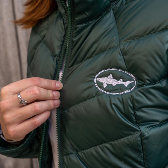 A person wearing the Patagonia W Dark Green 3-in-1 Parka with a white fish logo gently holds the zipper. The recycled down parka is paired with a fish-shaped ring on their index finger.