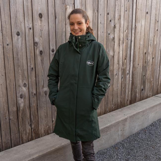 A person stands outdoors before a wooden fence, smiling and wearing the Patagonia W Dark Green 3-in-1 Parka with hands in the pockets.