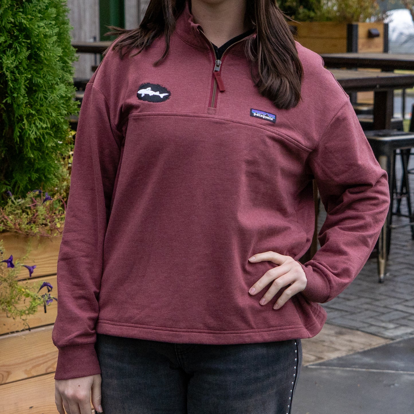 A person wears a Patagonia W Deep Red Ahnya Quarter Zip Pullover outdoors, standing with one hand on their hip near wooden planters and patio seating. The top of their head is partially out of frame.