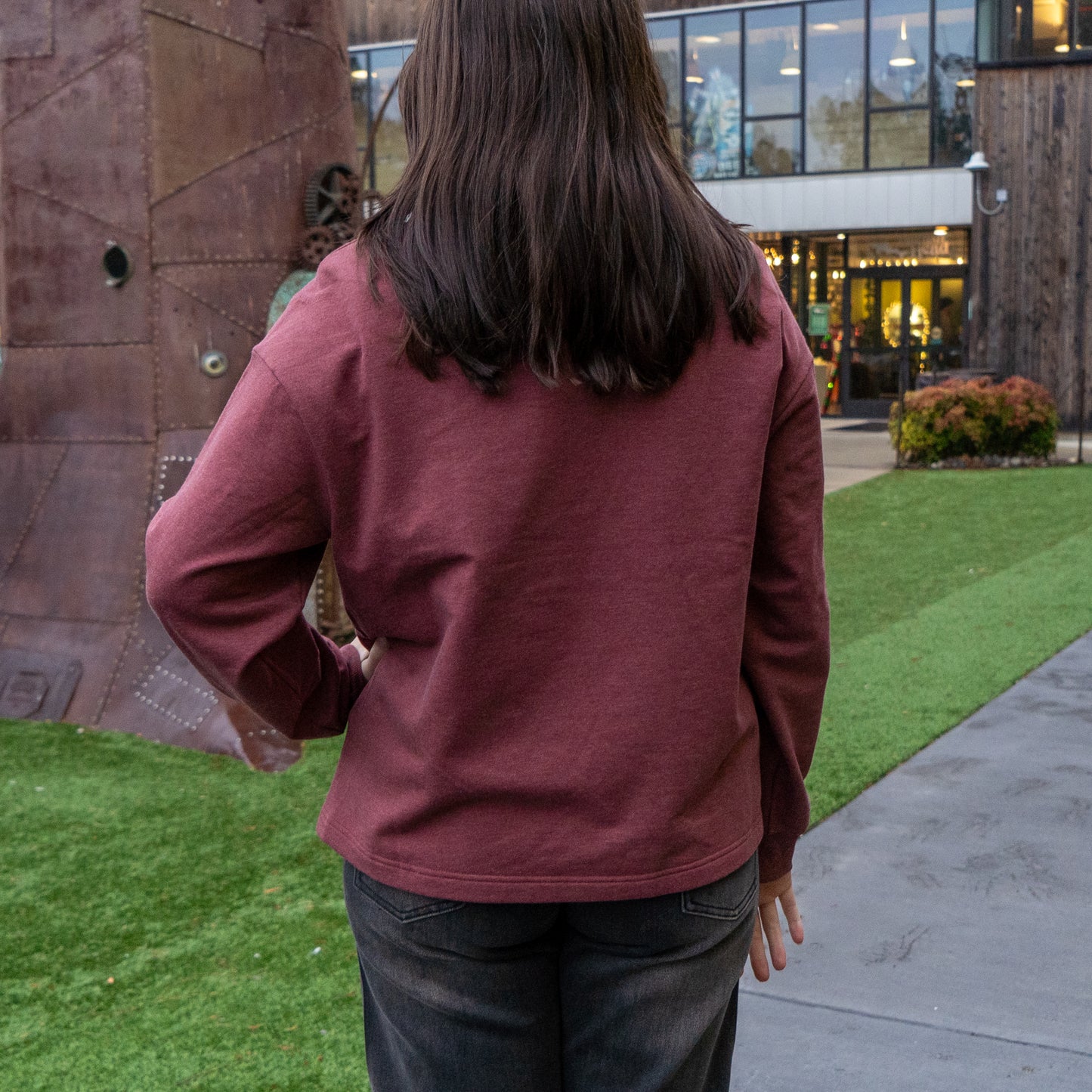 A person with long brown hair, wearing the Patagonia W Deep Red Ahnya Quarter Zip Pullover and dark jeans, stands on grass facing away from the camera near a modern building and large metal structure.