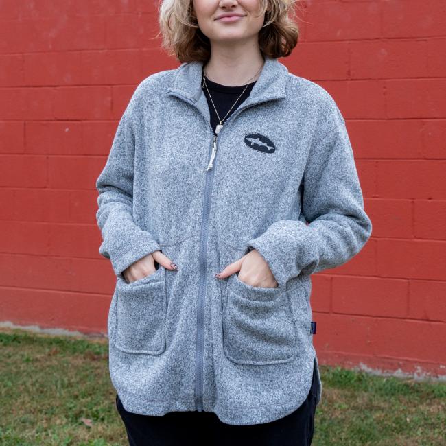 A person in a Patagonia W Heather Gray Full Zip Sweater Coat, made of recycled polyester fleece, stands before a red brick wall with hands in large front pockets and short curly hair showing.