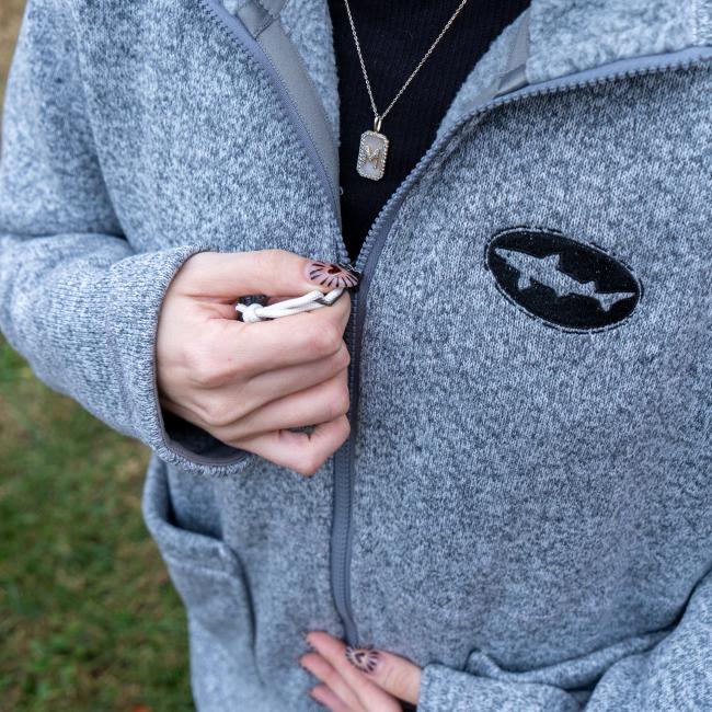 A person with patterned nails zips up a Patagonia W Heather Gray Full Zip Sweater Coat, standing outdoors on grass. They wear a necklace over a black top, and the sweater features recycled materials and Patagonia branding.