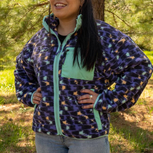 A person with long dark hair smiles outdoors, wearing the Patagonia W Purple Pattern Zip Jacket by Patagonia. The jacket features a zippered chest pocket and light green accents. Trees and grass are visible in the background.