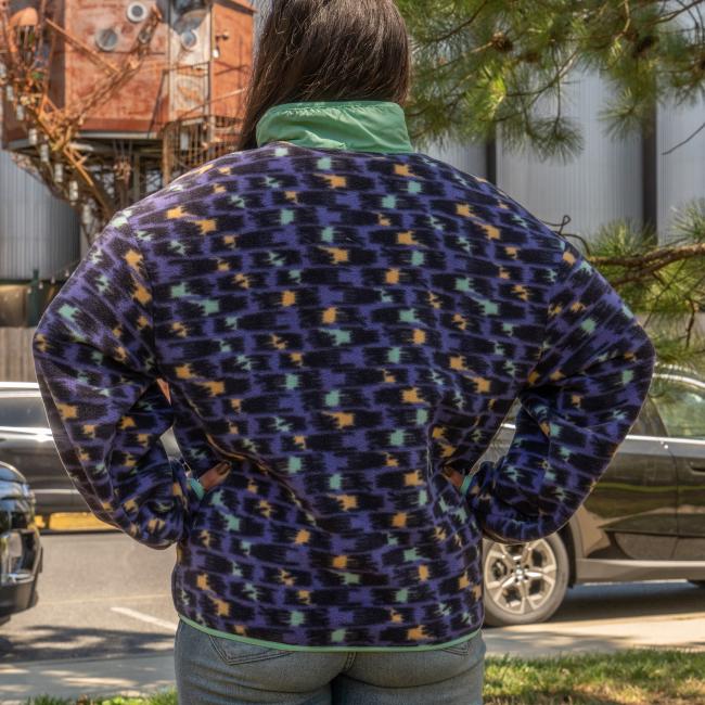 A person stands outdoors with their back to the camera, wearing a Patagonia W Purple Pattern Zip Jacket. Trees, a black car, and an industrial building are visible in the background.