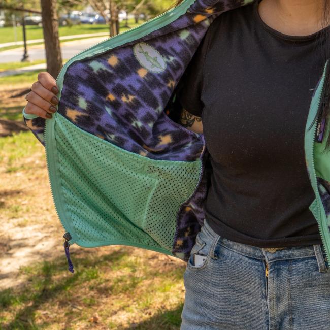 A person outdoors holds open the Patagonia W Purple Pattern Zip Jacket by Patagonia, showing its Fair Trade Certified recycled polyester shell, patterned purple, black, and yellow lining, and mesh inner pocket with grass and trees behind.