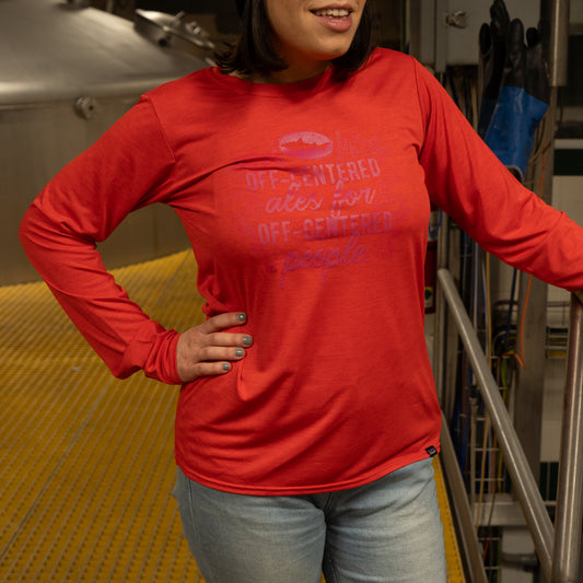 A person wears a Patagonia W Red Long Sleeve Runner by Patagonia with light jeans, standing indoors on a yellow metal floor, one hand on their hip and the other on a railing. The shirt has text printed on the front.