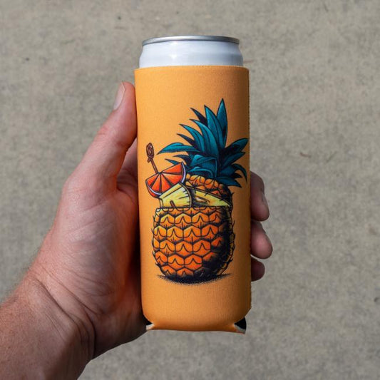 A hand holds a tall can wrapped in the Merchtank Peach Pineapple Slim Can Cooler, featuring a pineapple drink illustration with orange, straw, and cocktail umbrella against a plain background.