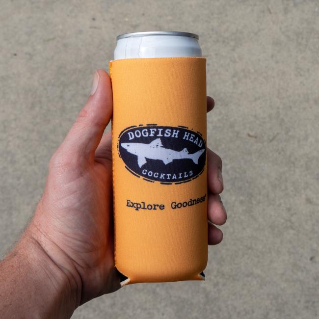 A hand holds a can wrapped in an orange Merchtank Peach Pineapple Slim Can Cooler on a plain, light-colored background.