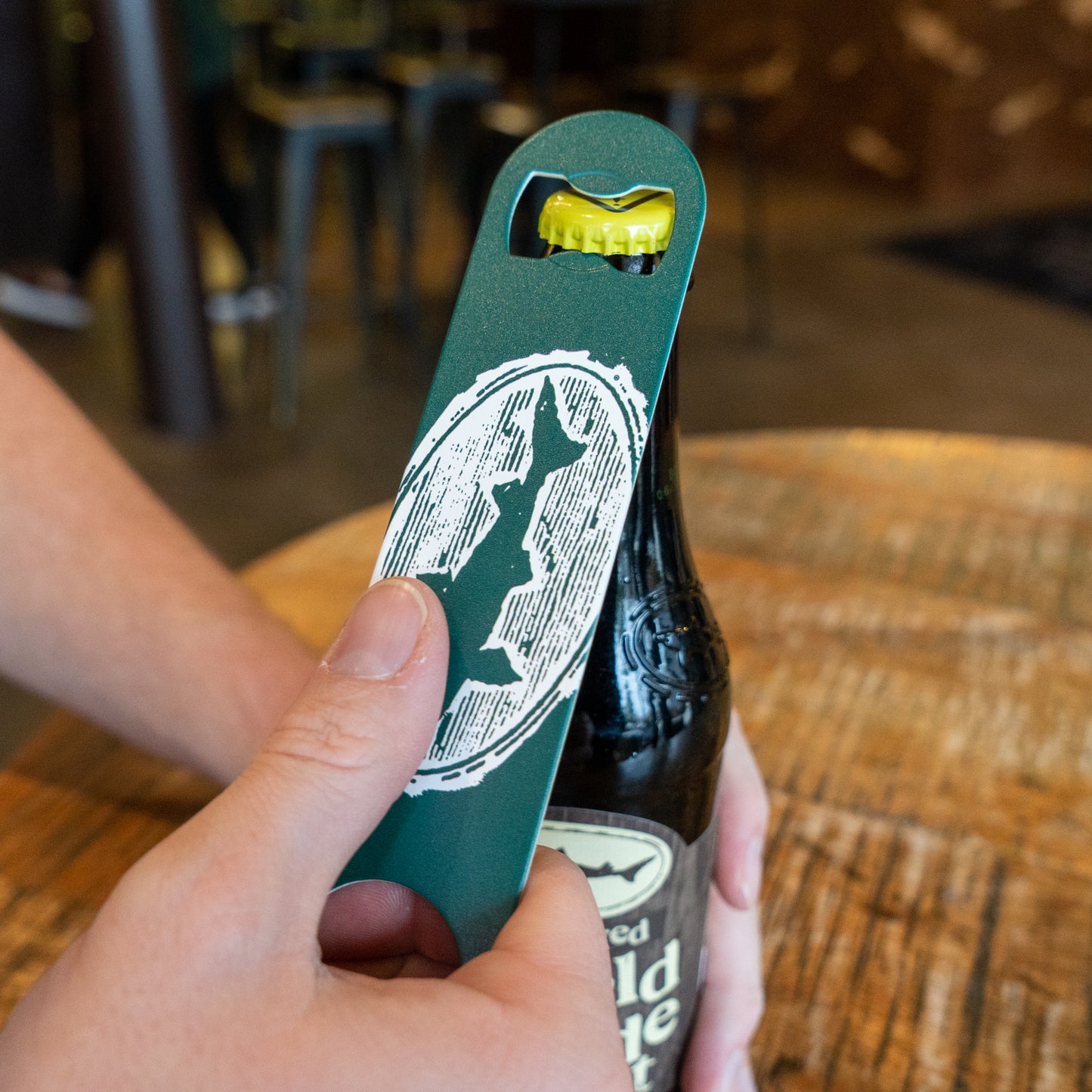 A person uses the Merchtank Bartender Opener, featuring a green design with a white logo, to open a bottled beverage on a wooden table.