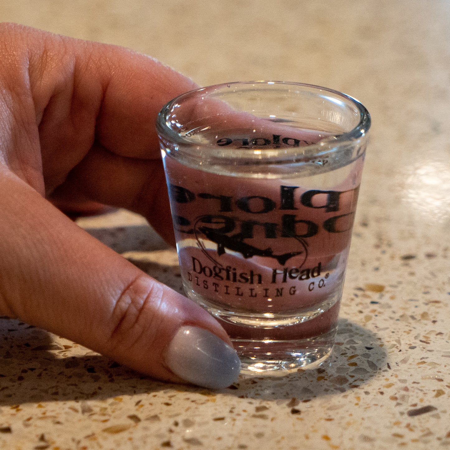 A hand with light blue painted nails holds a Grandstand Glassware & Apparel Shot Glass (2.5 oz), filled with clear liquid, on a speckled countertop.