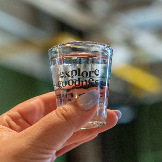 A hand with light purple nail polish holds a clear Grandstand Glassware & Apparel Shot Glass filled with spirits, featuring the words "explore goodness." The background is blurred.