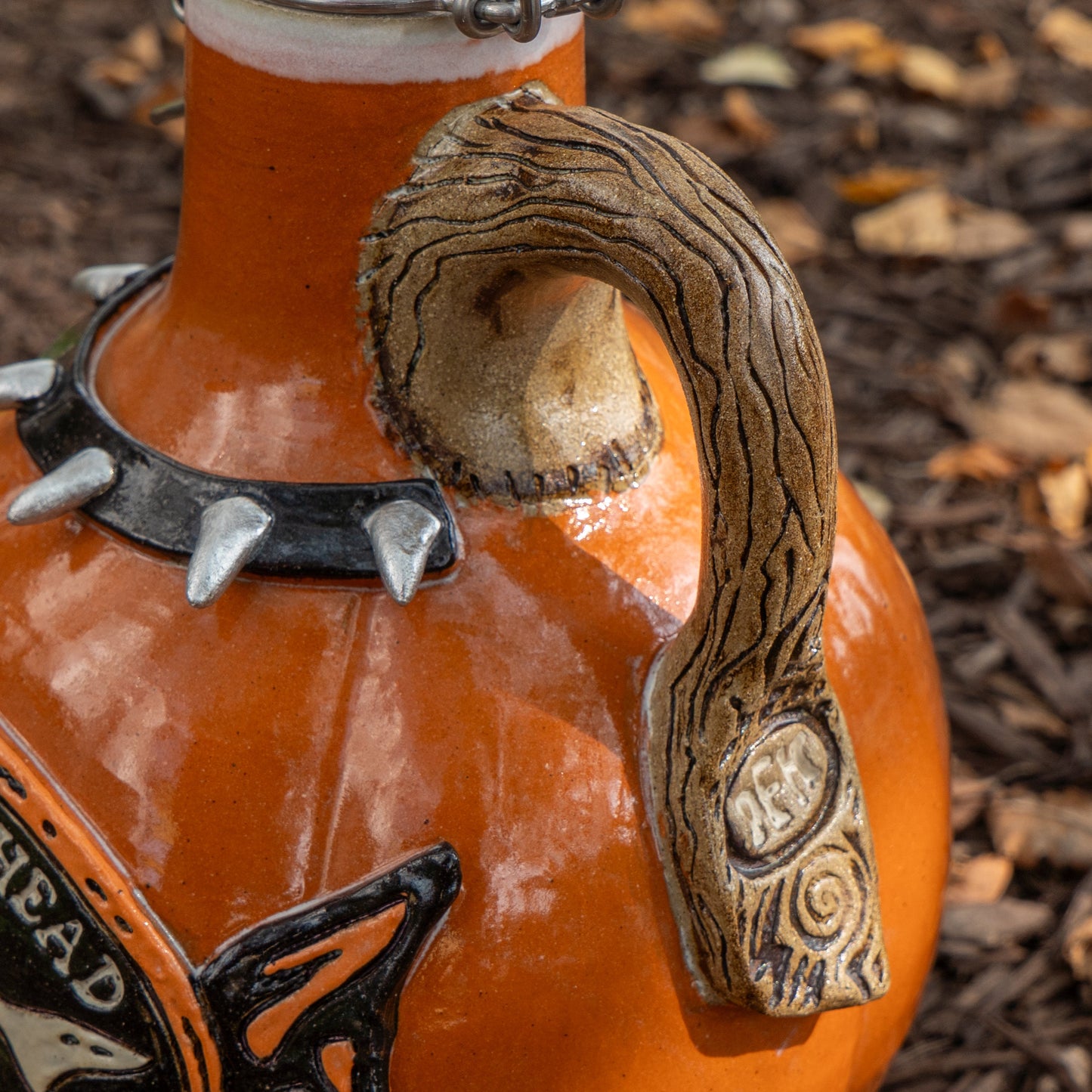Close-up of Romanick Pottery's Punk Punkin Ceramic Growler, featuring orange glaze, a wood-like handle, spiked collar, and swirl details. Shown on brown mulch with scattered leaves—an ideal piece for limited edition growler collectors.