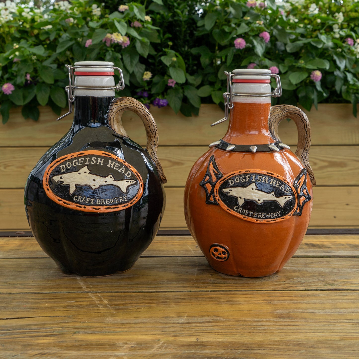 Two limited edition Romanick Pottery Punk Punkin Ceramic Growlers, one black and one orange, are side by side on a wooden surface with green plants and pink flowers in the background.