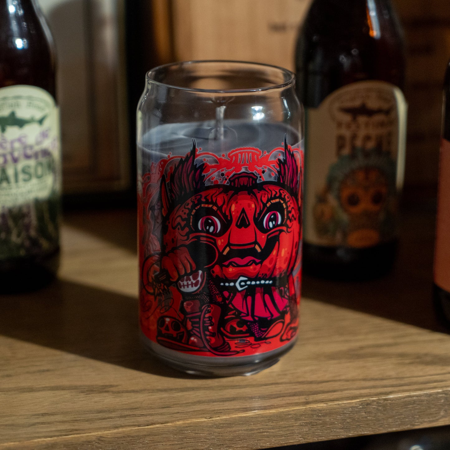 A clear glass featuring a bold red and black demon face sits on a shelf alongside brown beer bottles and the Punkin Can Glass Candle from Unwined Candles, LLC. Warm light highlights the candle, glass, and surrounding surfaces.